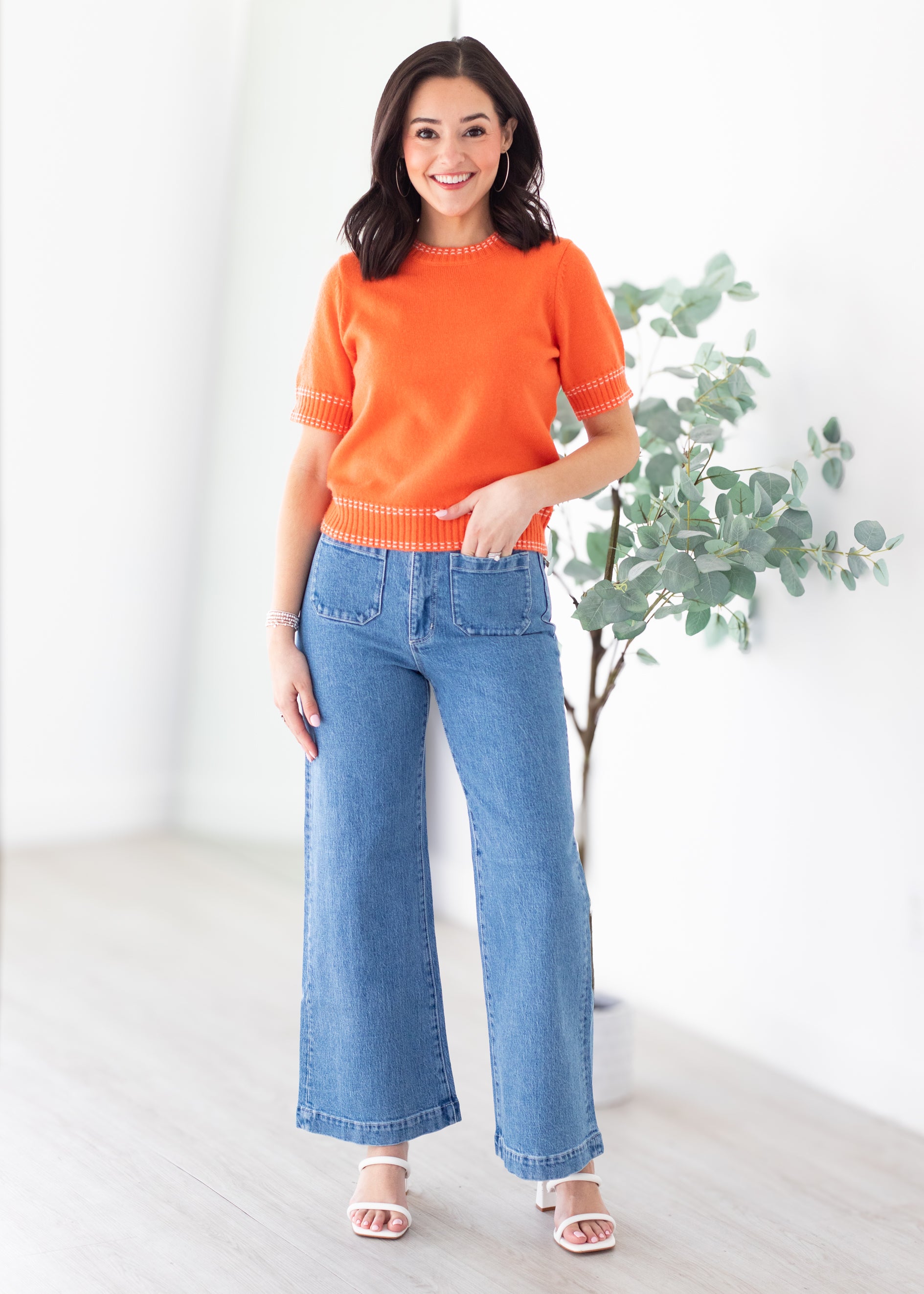 Woman wearing an orange sweater and blue jeans standing in a bright room with a plant.
