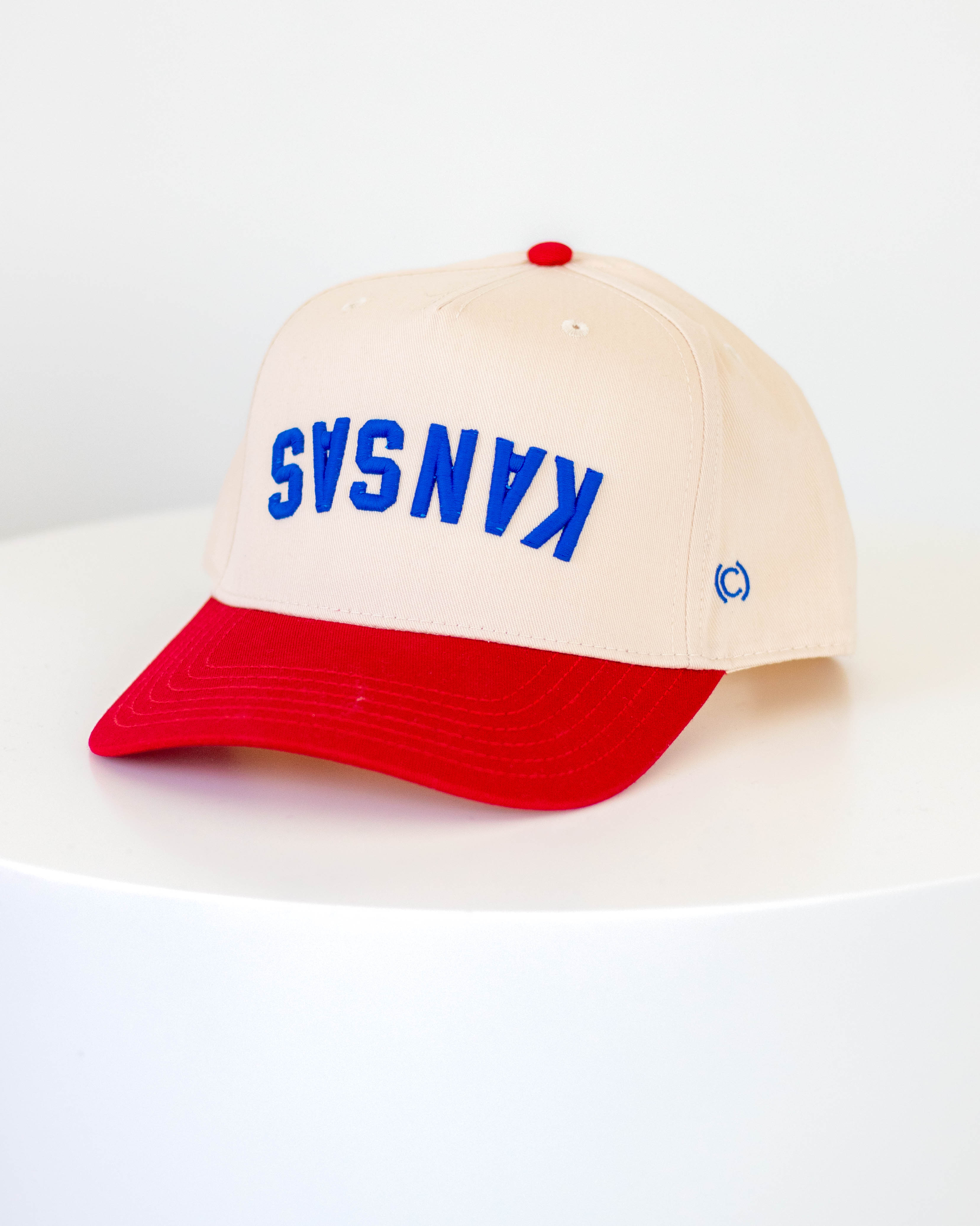 Red and cream colored hat with "KANSAS" embroidered upside down in blue lettering on a white background