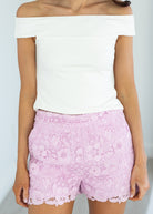 Person wearing a white off-shoulder top and pink lace shorts on a plain background