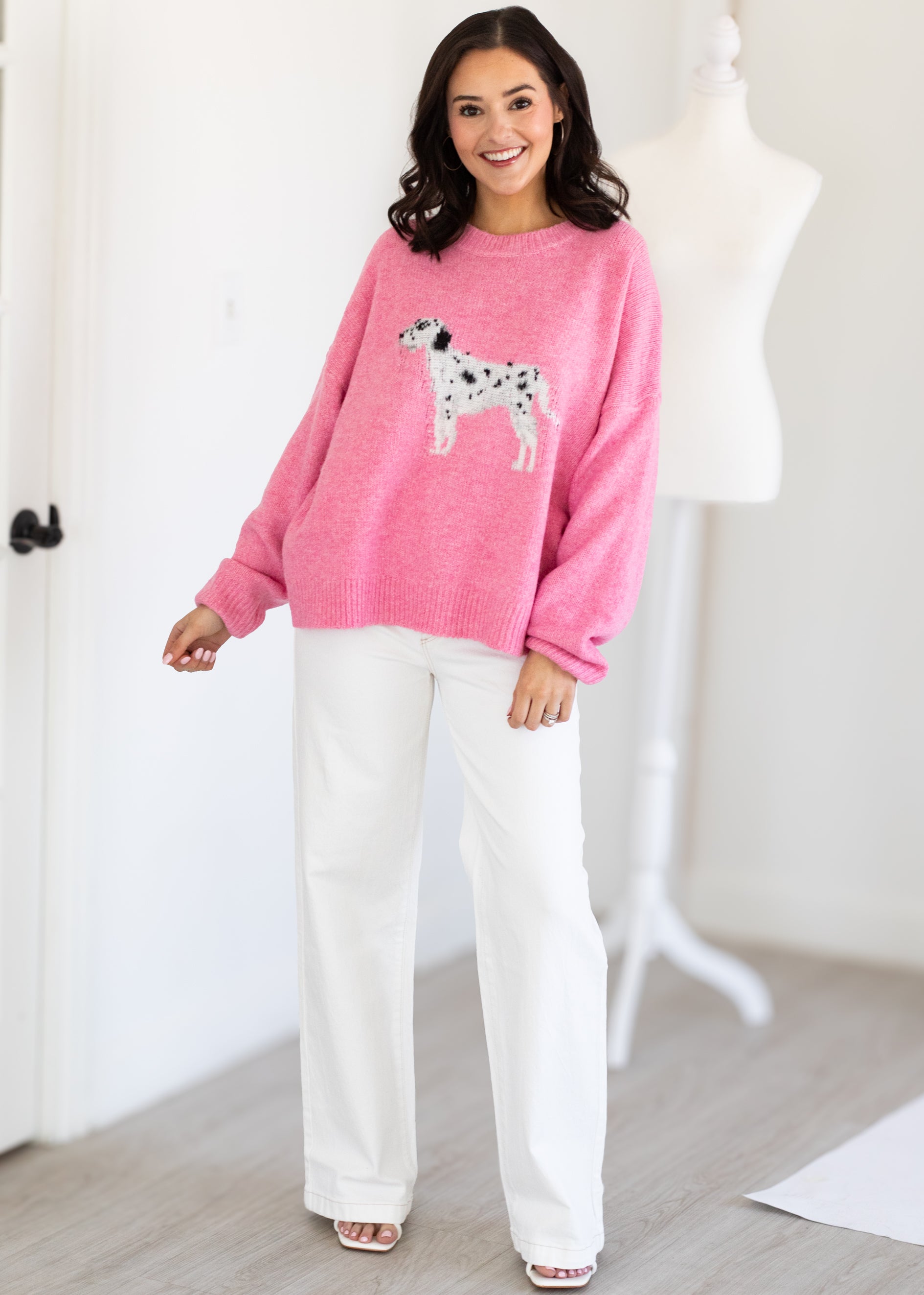 Woman wearing a pink sweatshirt with a dog graphic and white pants in a room.