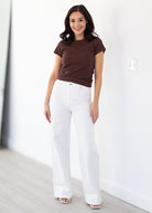 Woman wearing a brown t-shirt and white pants standing against a white wall.