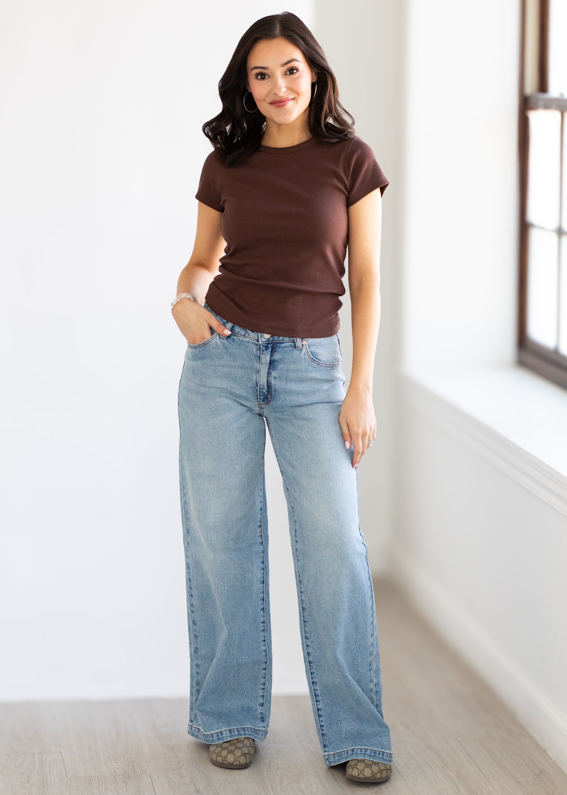 Woman wearing a brown t-shirt and light blue jeans standing in a room with a window.