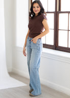 Woman wearing a brown top and blue jeans standing in a bright room with a window.
