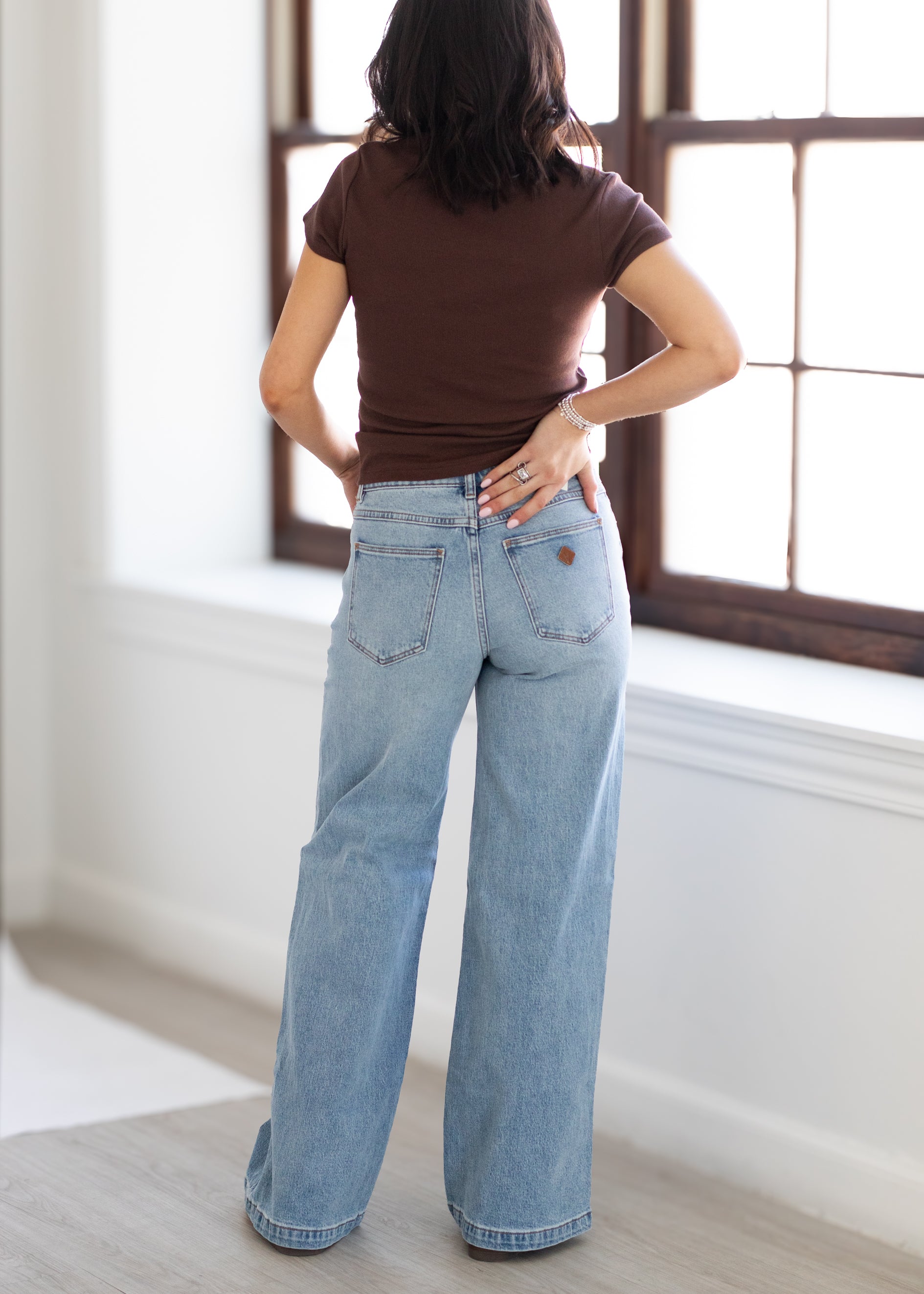 Person wearing a brown top and light blue jeans standing in a room with large windows.