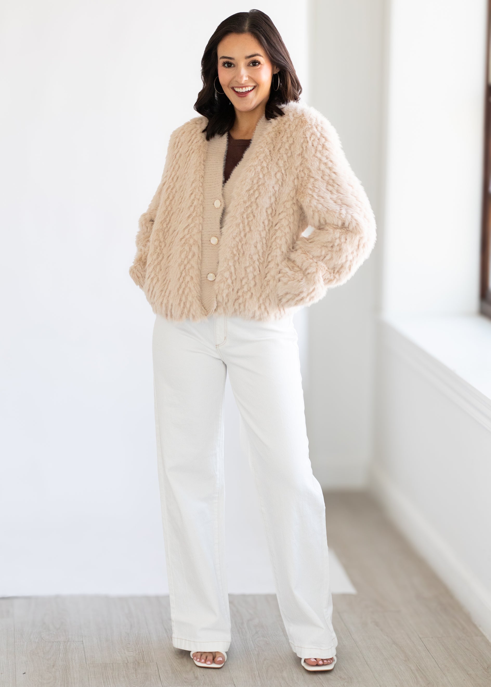 Woman wearing a beige knitted cardigan and white pants in a minimalistic room.