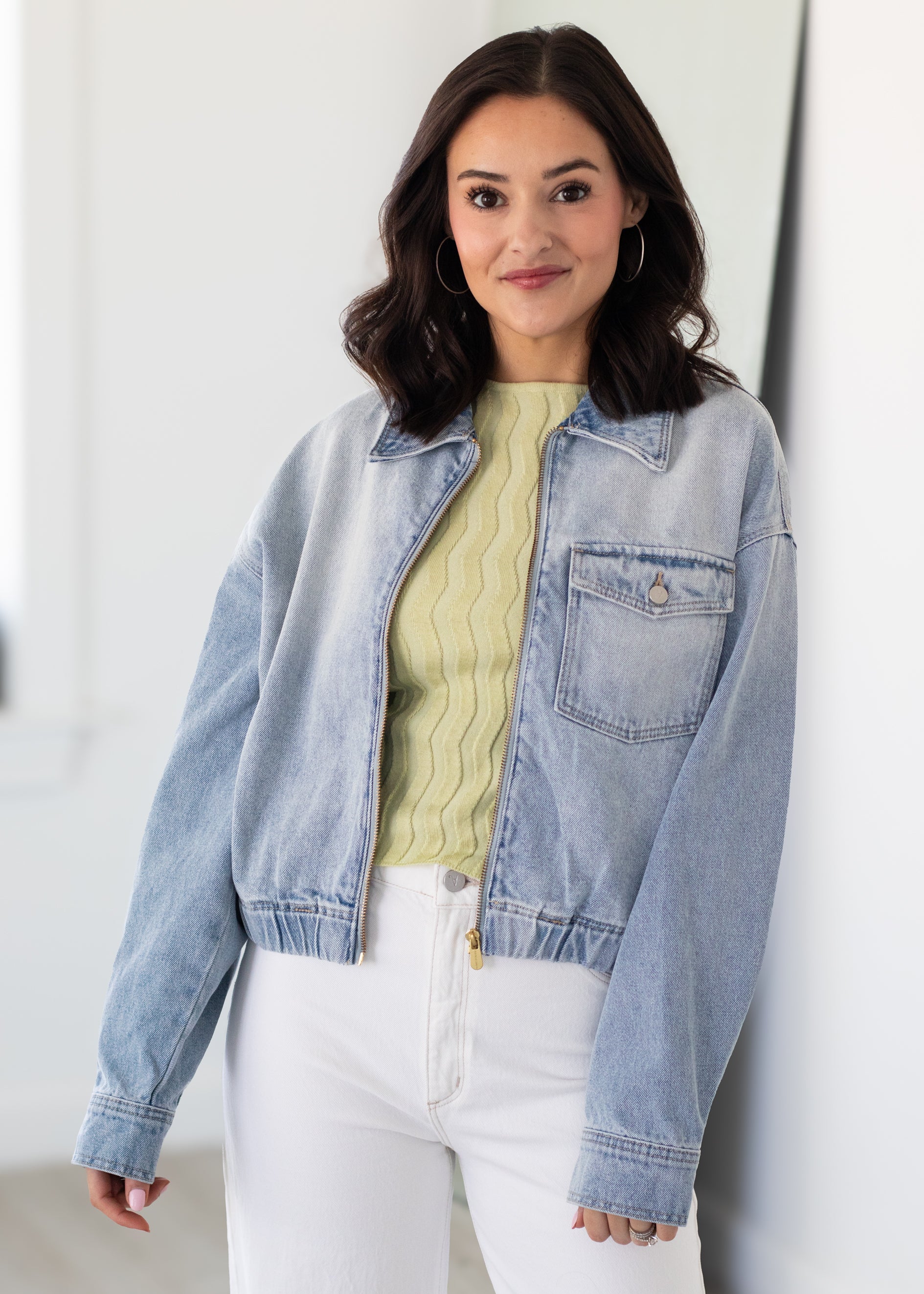 Woman wearing a light blue denim jacket over a yellow sweater and white pants against a white background