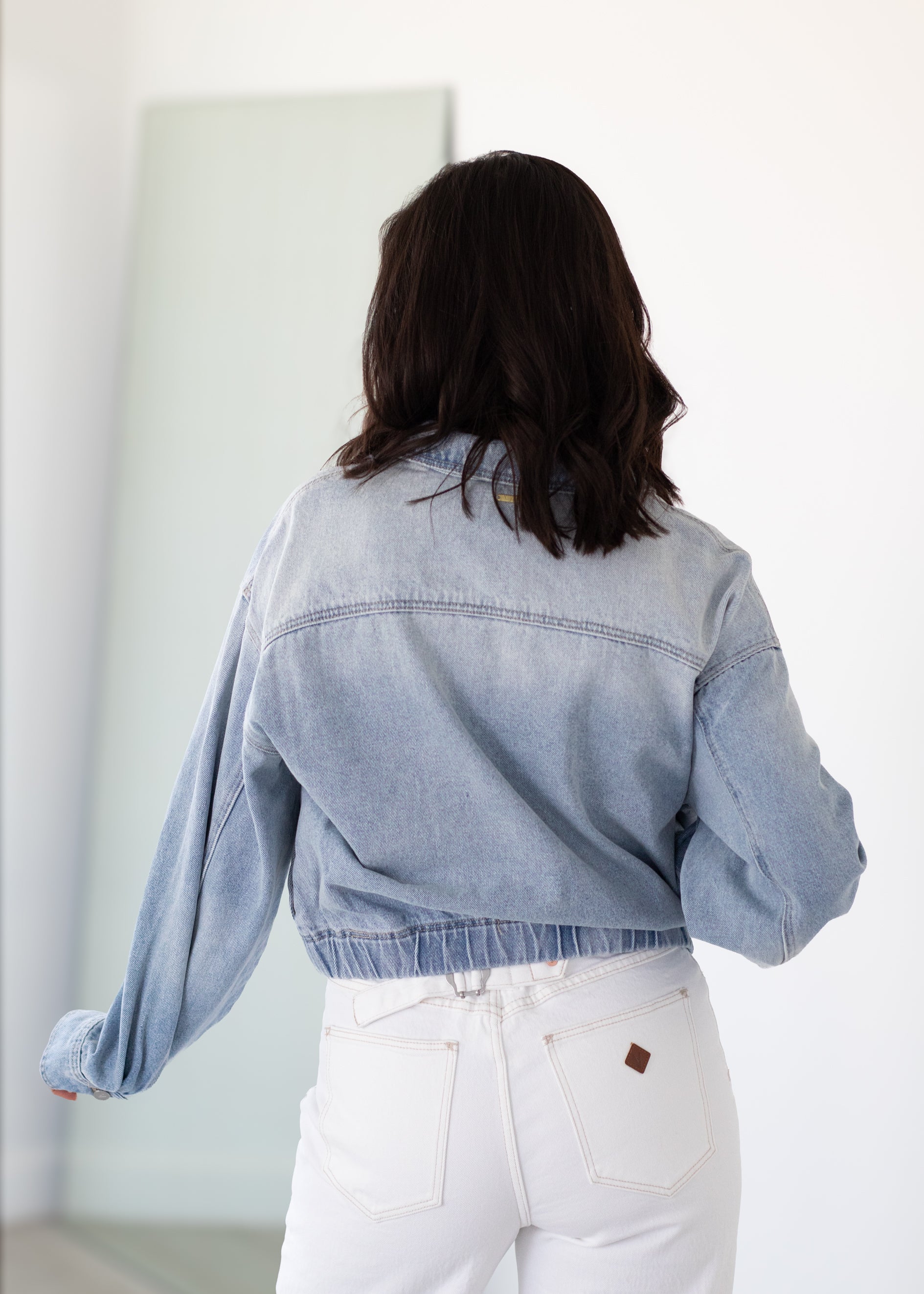 Person wearing a light blue denim jacket and white pants in a minimal indoor setting