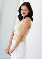 Woman wearing a light green sleeveless top and white pants against a white background