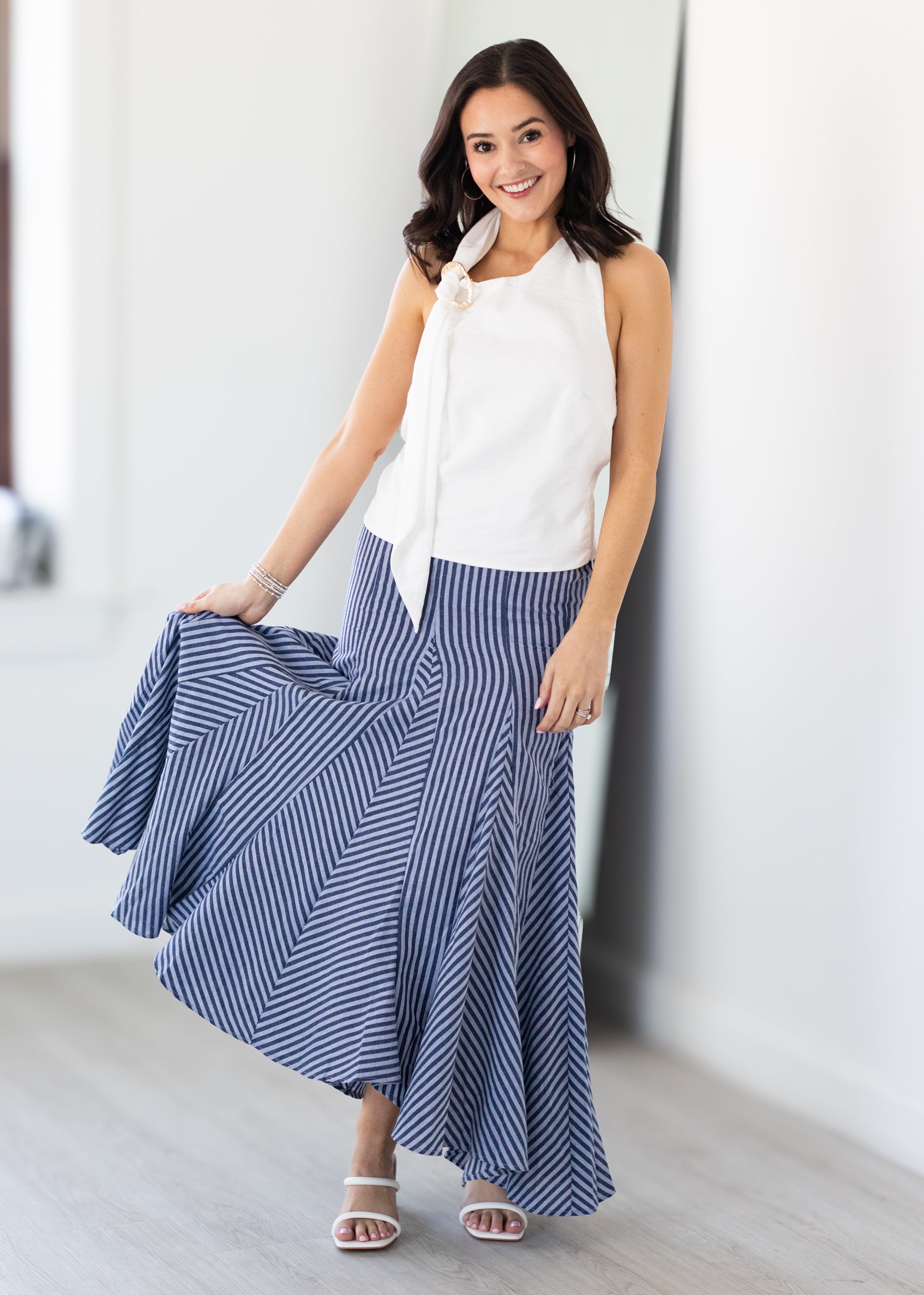 Woman wearing a white sleeveless top and blue striped skirt in a minimal indoor setting.