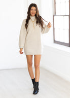 Woman wearing a beige sweater dress with black boots in a bright room.