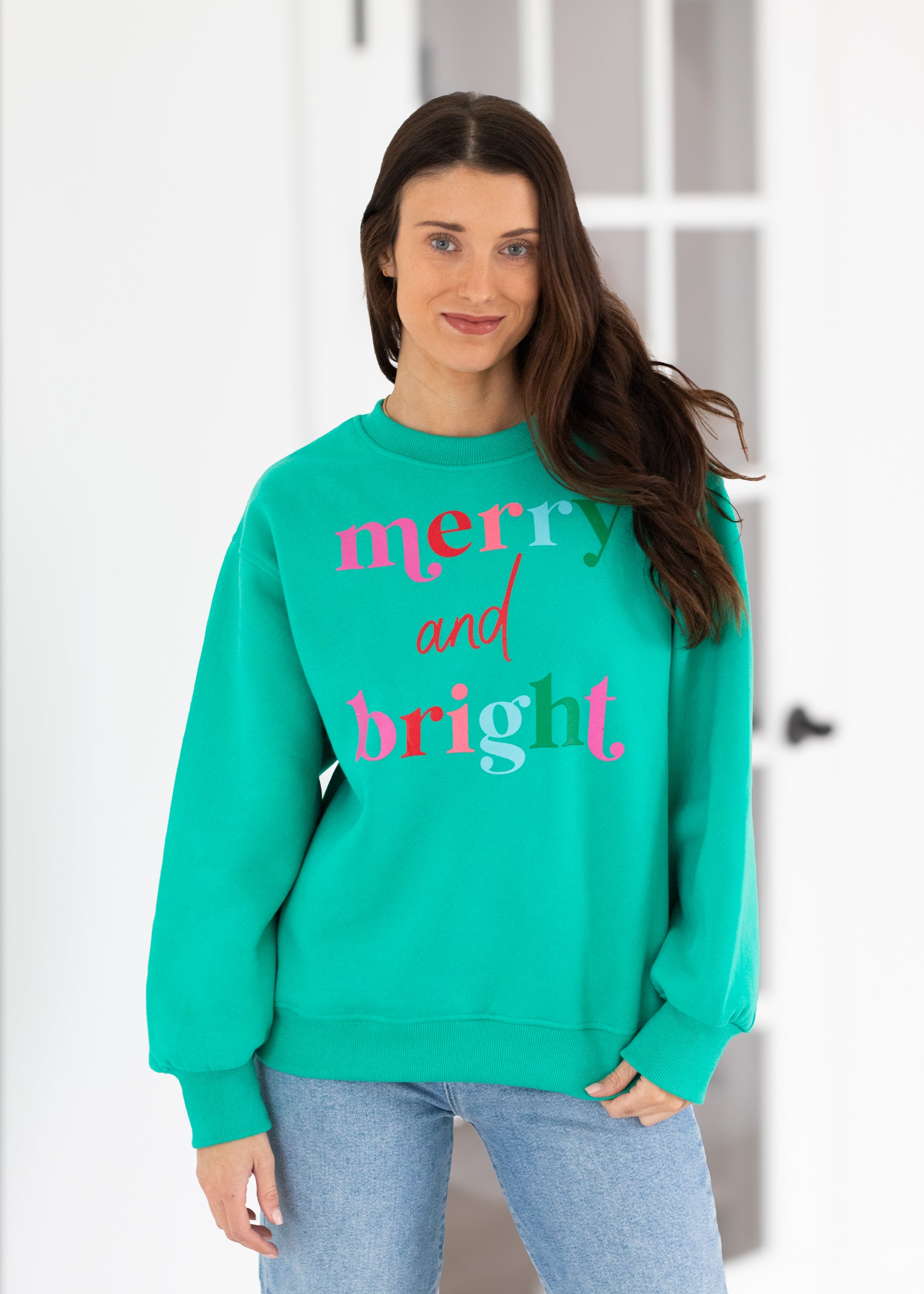 Woman wearing a teal sweatshirt with 'merry and bright' text, standing against a white wall.