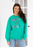 Woman wearing a teal sweatshirt with 'merry and bright' text, standing against a white wall.
