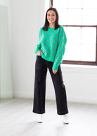 Woman wearing a green sweater with pink patterns and black pants in a bright room.