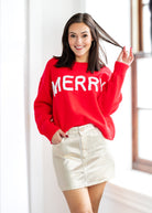 Woman wearing a red 'MERRY' sweater and gold skirt indoors.