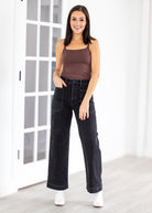 Woman wearing a brown tank top and black jeans standing in a room with white walls and a glass door.