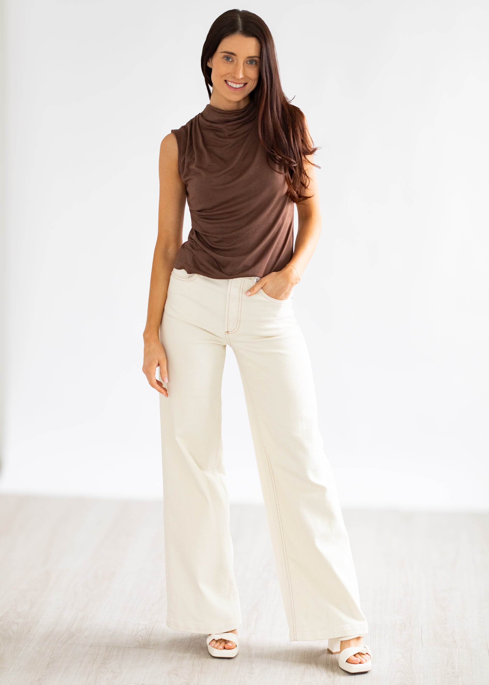 Woman wearing a brown sleeveless top and white pants on a plain background