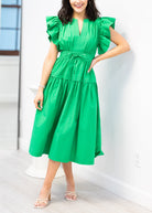 Woman wearing a green dress standing indoors with a white wall and light-colored floor.