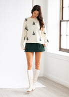 Woman wearing a cream sweater with tree patterns, green skirt, and white knee-high boots in a bright room.