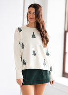 Woman wearing a white sweater with green Christmas tree patterns in a minimal indoor setting.