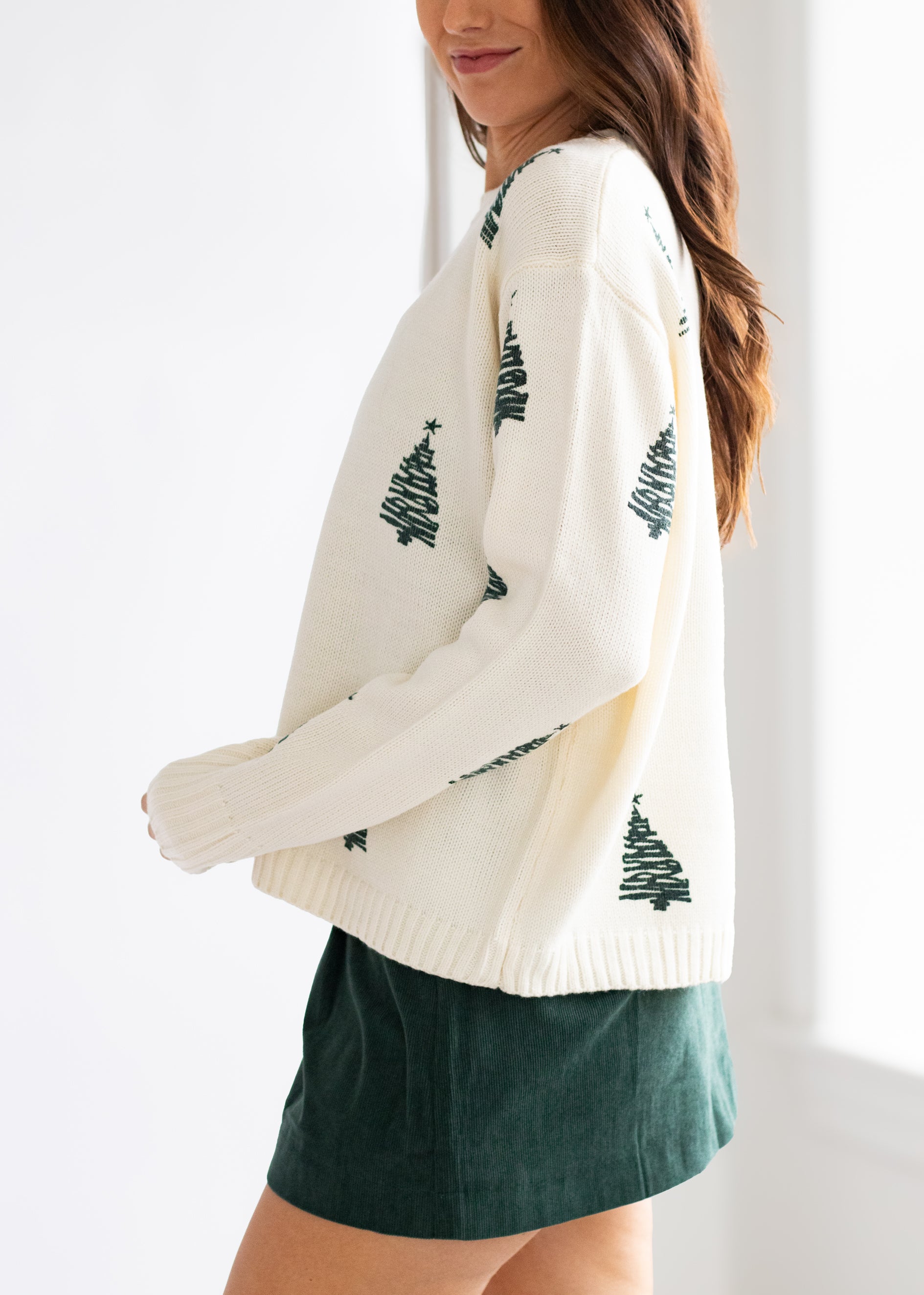 Woman wearing a cream sweater with tree patterns and green shorts on a white background