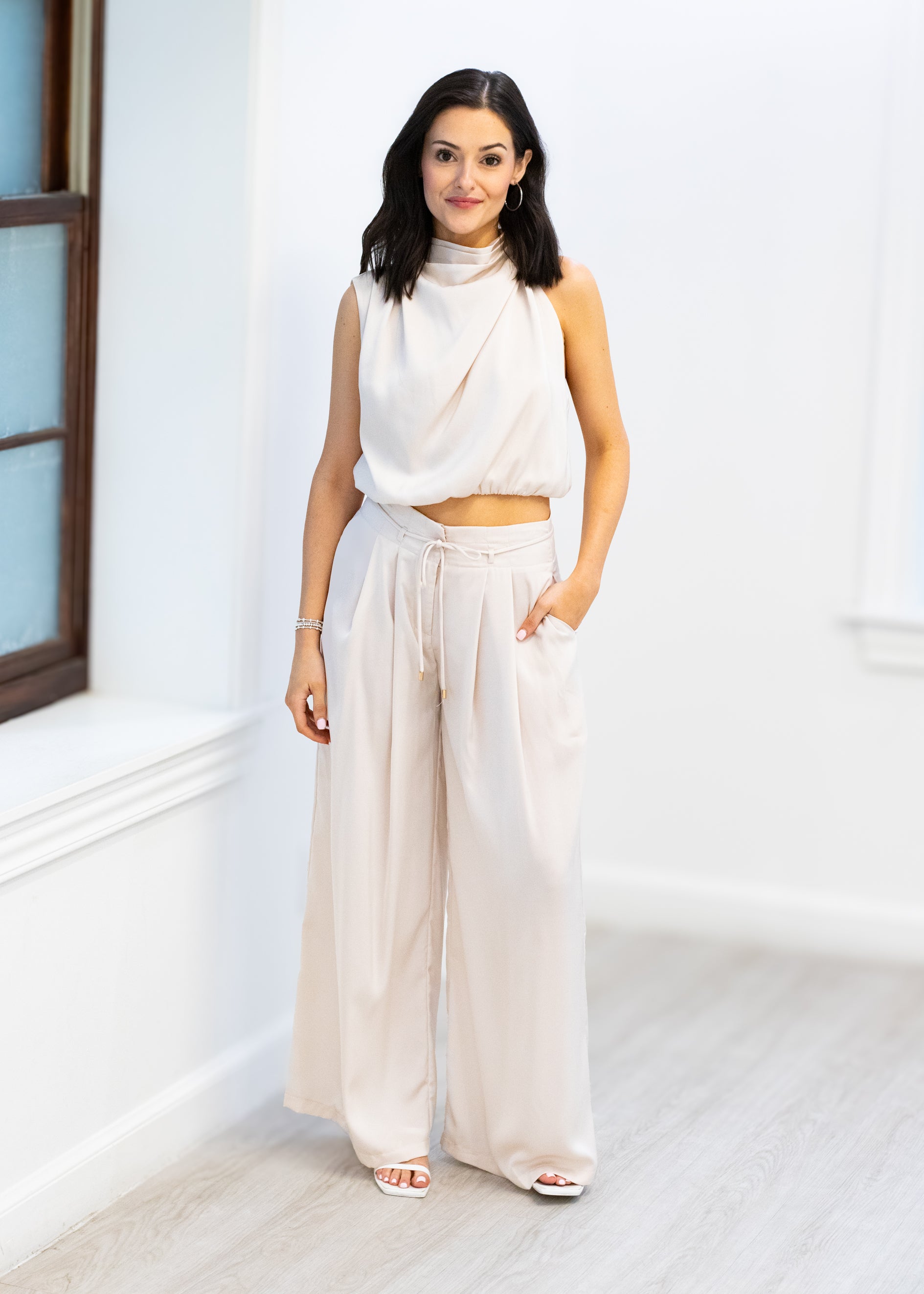 Woman wearing a white sleeveless top and beige wide-leg pants standing in a bright room.