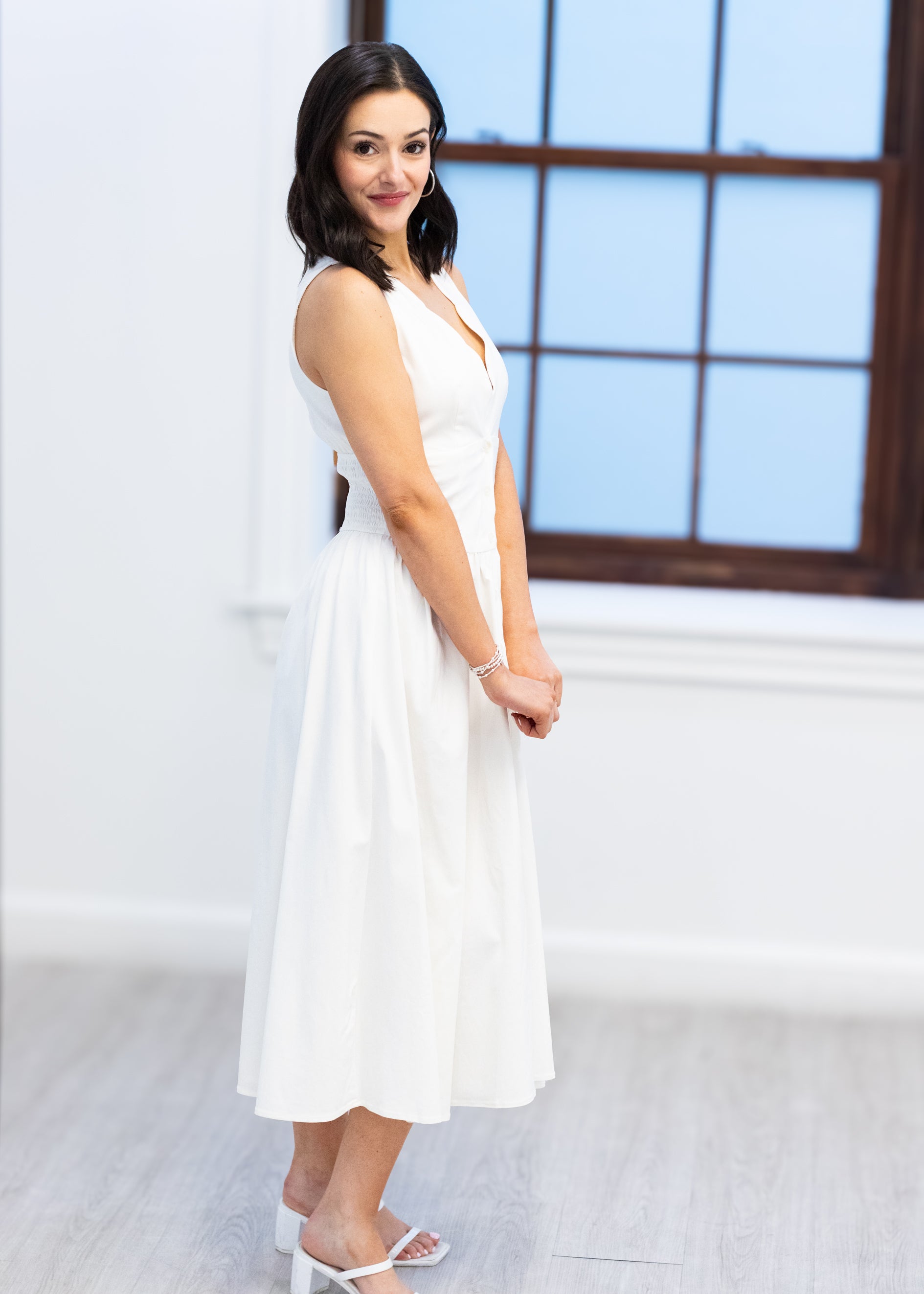 Woman in a white dress standing in a room with large windows.