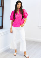 Woman wearing a bright pink blouse and white pants standing in a minimalistic room.
