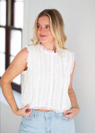 Woman wearing a white sleeveless top with fringe details and denim shorts indoors.