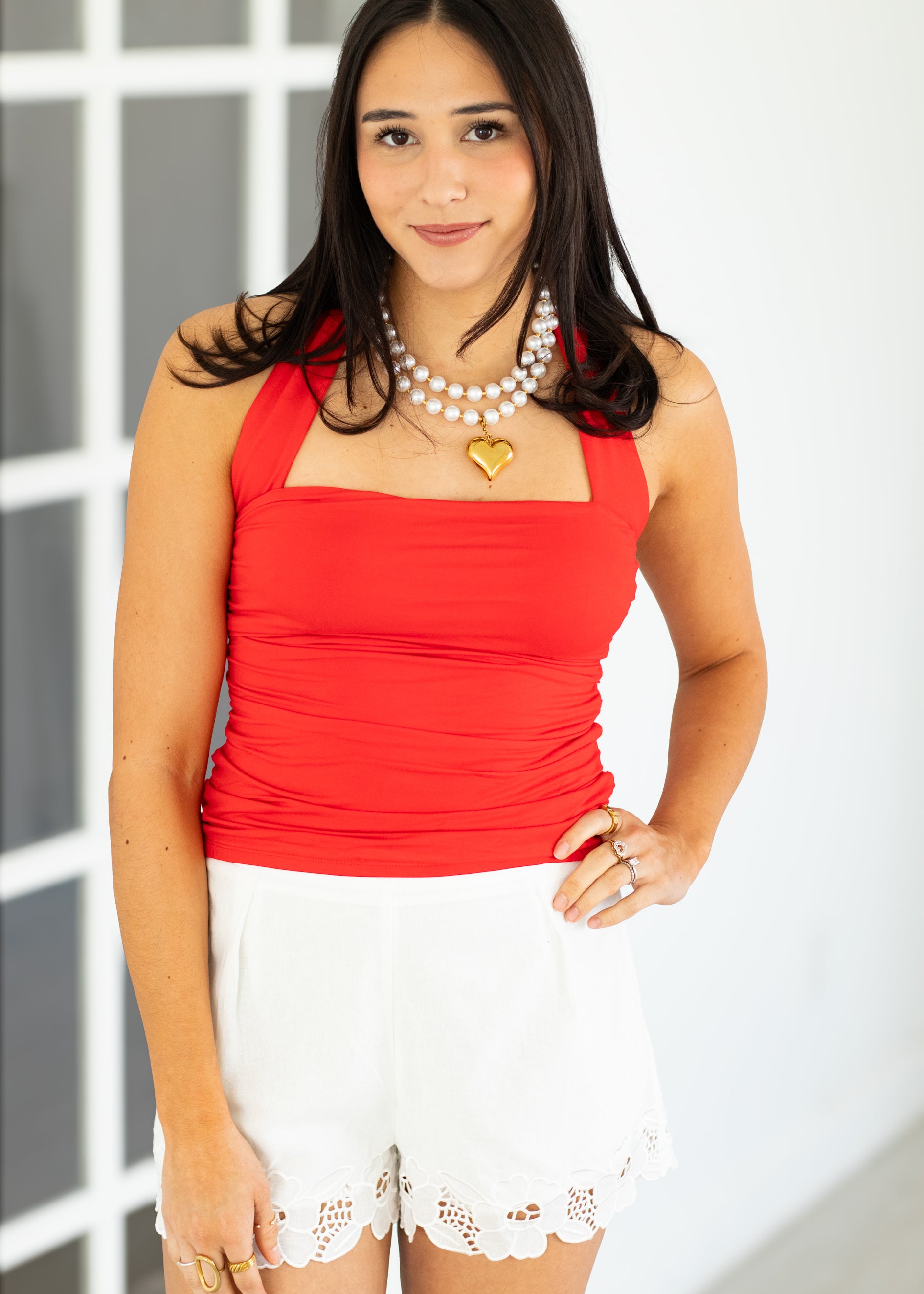 Woman wearing a red top and white shorts with a pearl necklace.