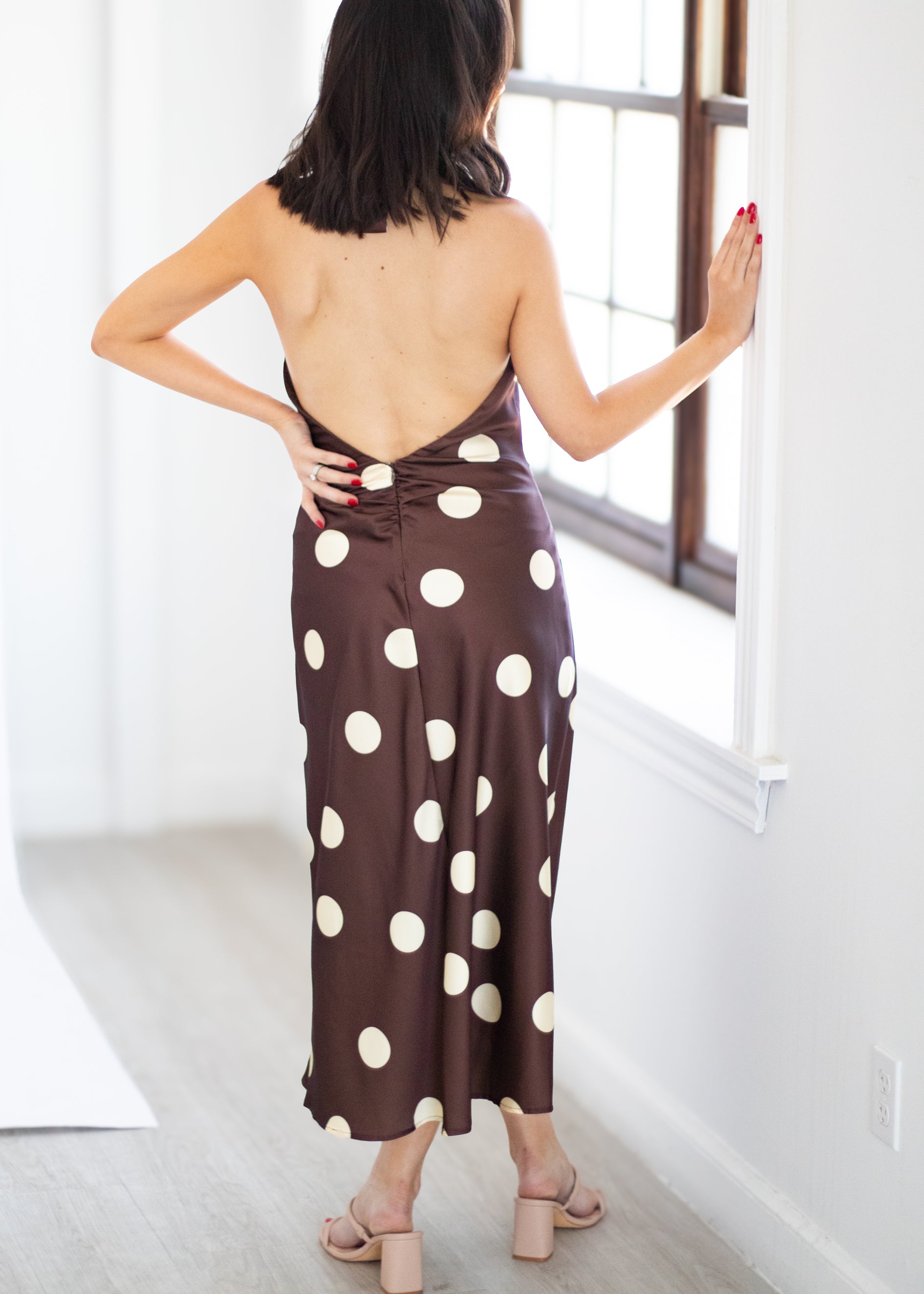 A woman stands with her back to the camera wearing a brown satin-like maxi dress with large white polka dots and an open back, posed in a bright room next to a window.