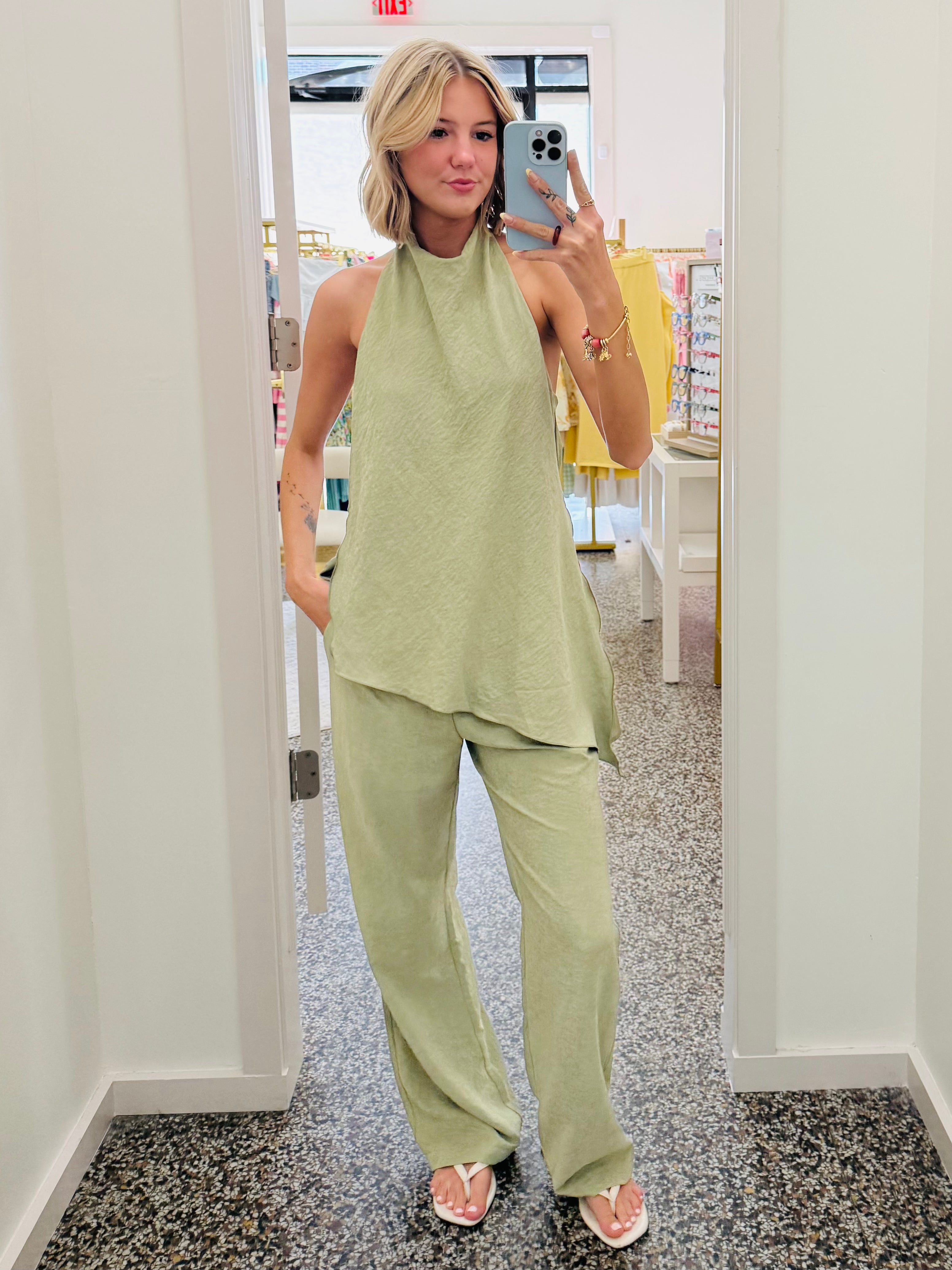 A woman takes a mirror selfie in a boutique wearing a light sage green two-piece lounge set consisting of a sleeveless high-neck top and matching wide-leg pants.