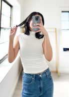Person taking a mirror selfie wearing a white turtleneck and blue jeans in a bright room.