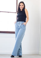 Woman wearing a black sleeveless top and light blue jeans standing against a white wall.
