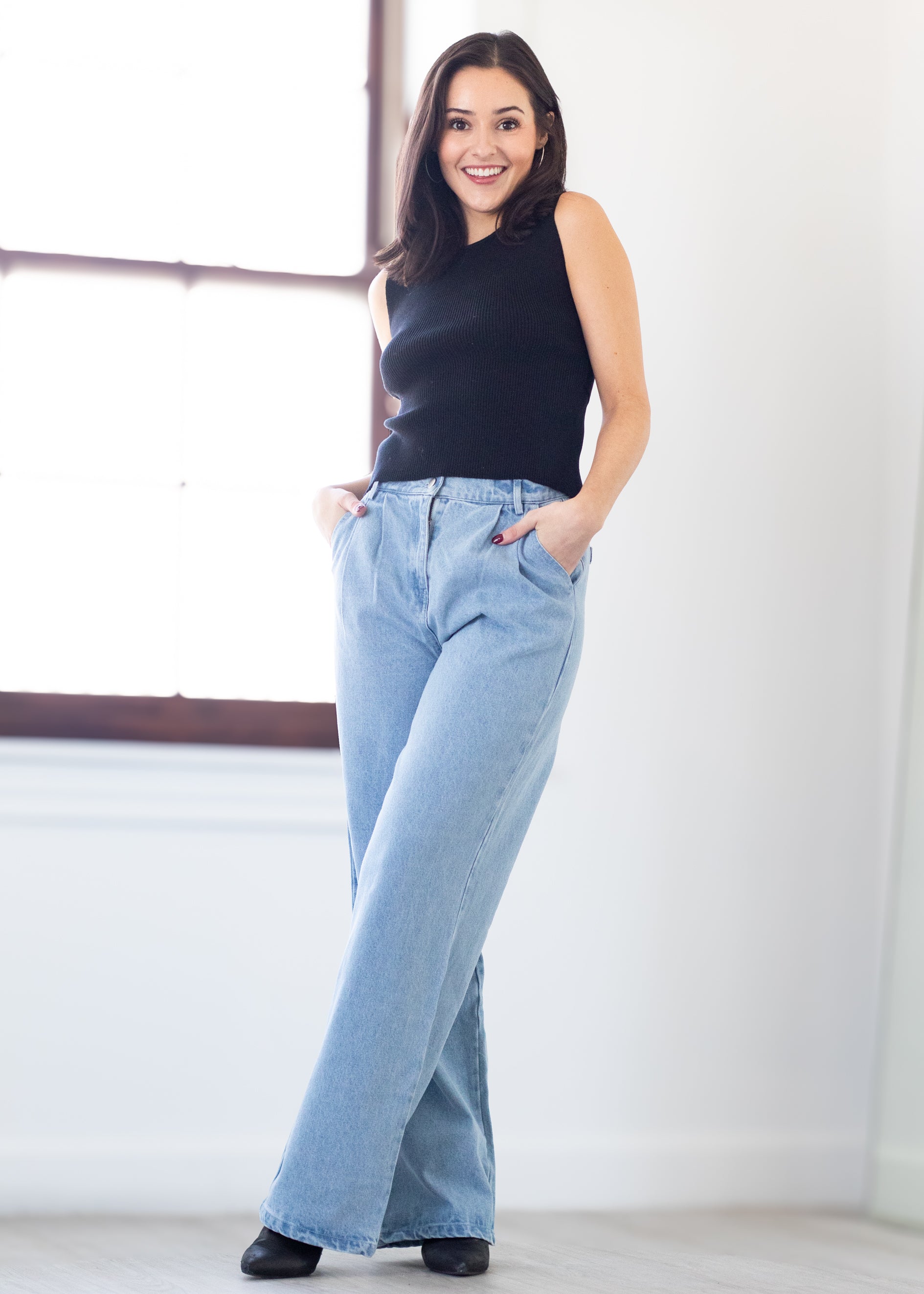 Woman wearing a black sleeveless top and light blue jeans standing against a white wall.