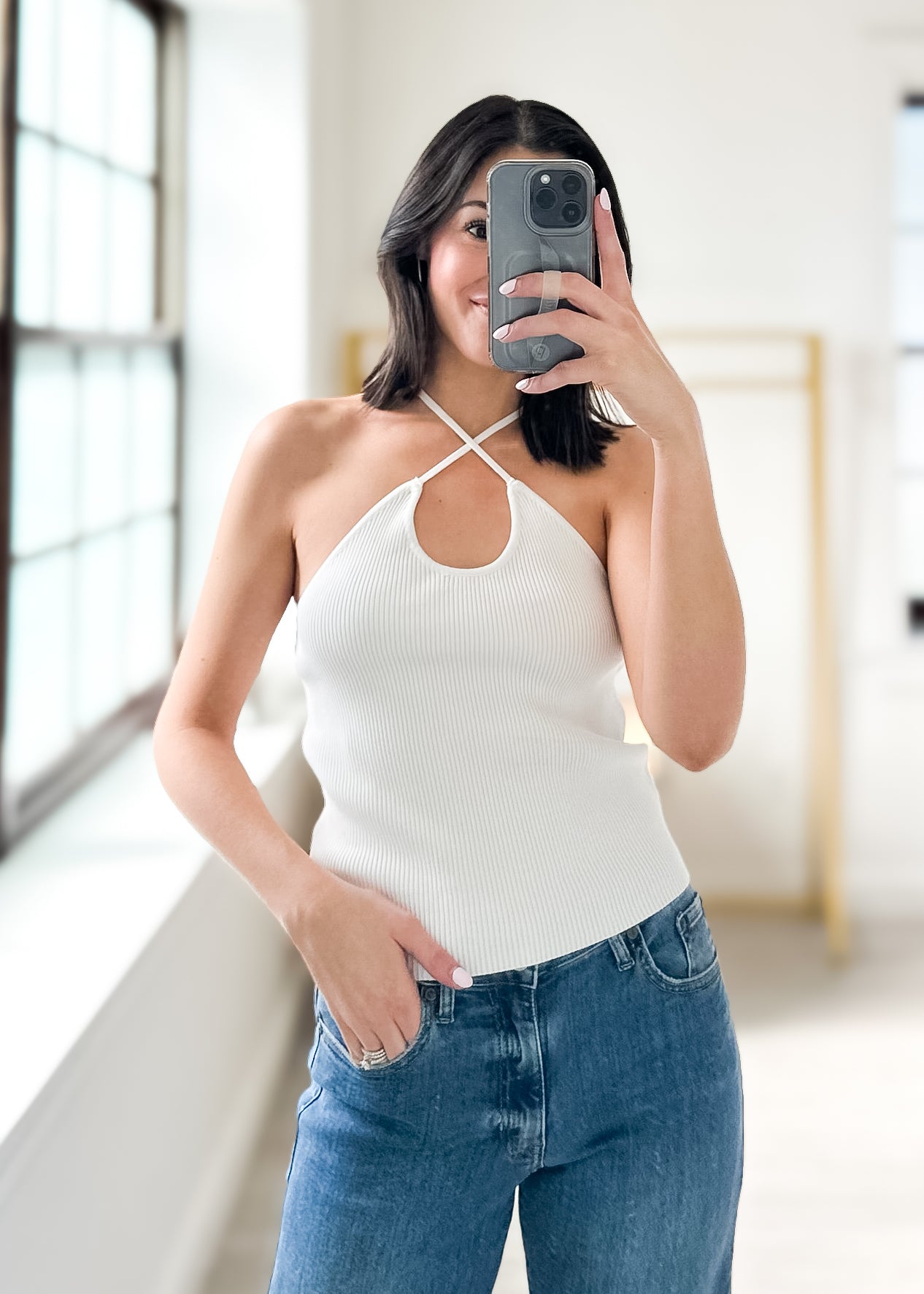 Woman taking a mirror selfie wearing a white halter top and blue jeans.