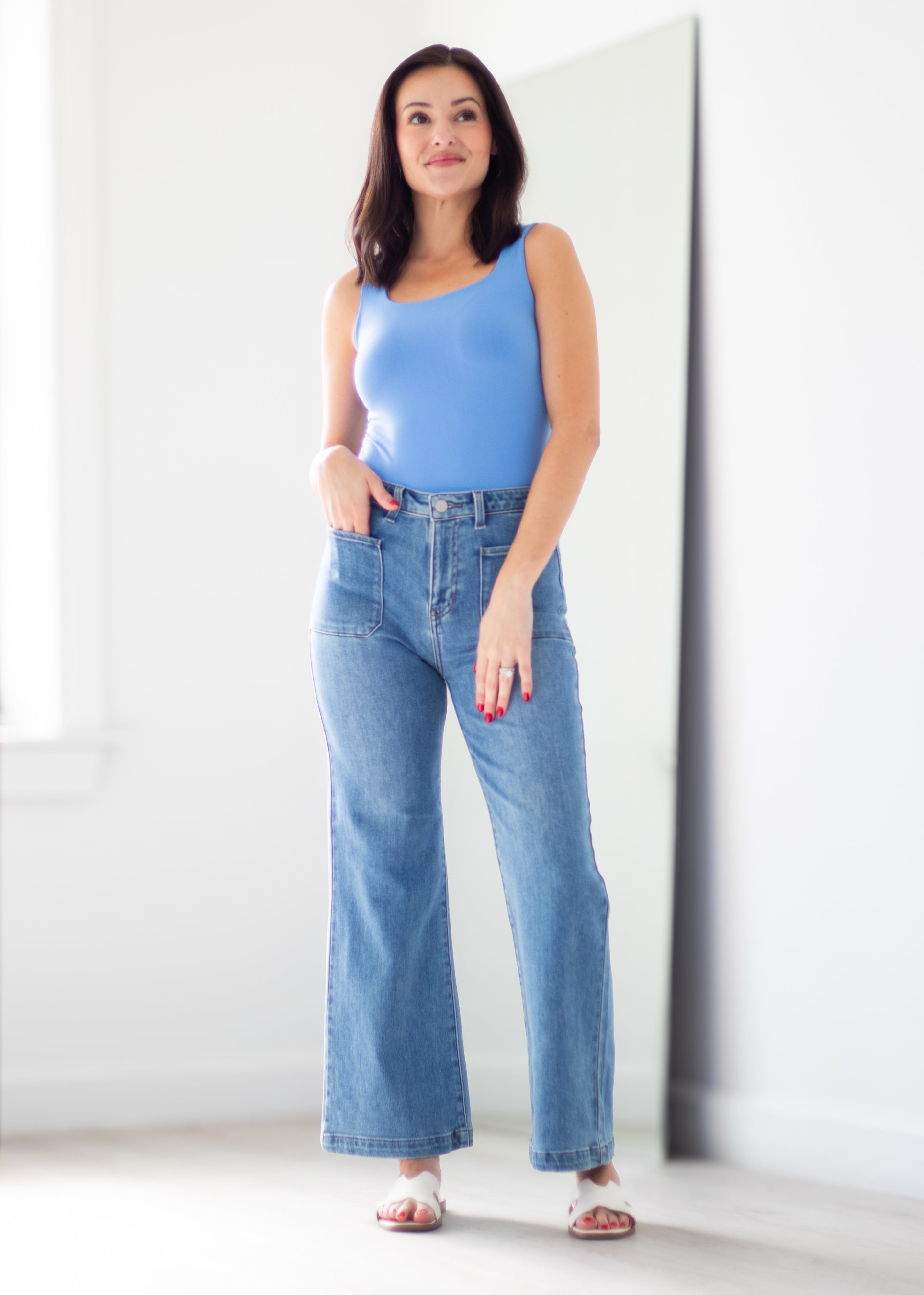 A woman modeling a light blue sleeveless top and high-waisted, wide-leg blue denim jeans in a bright, minimalist white room.