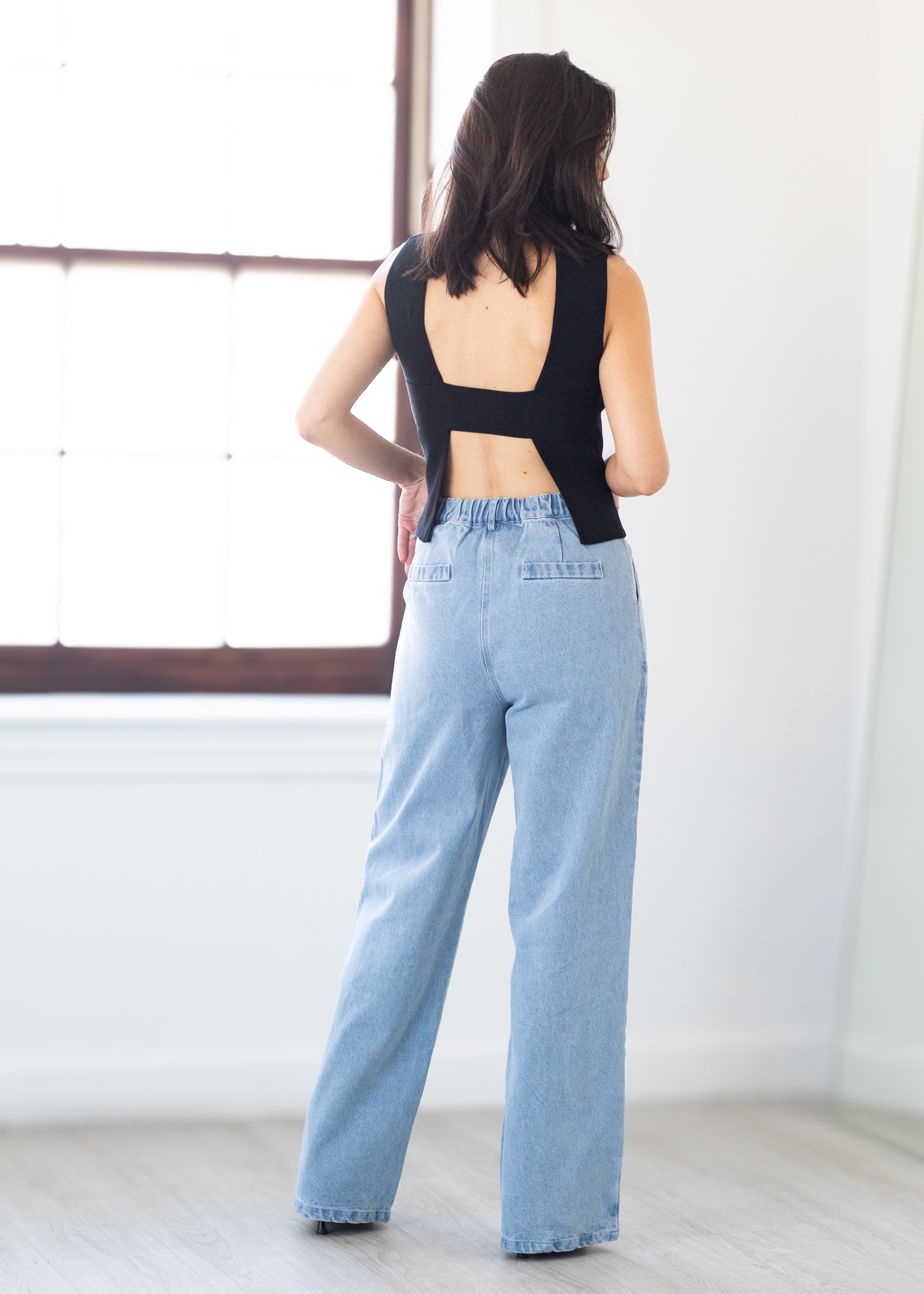 Person wearing a black sleeveless top and light blue jeans standing in a minimalistic room.
