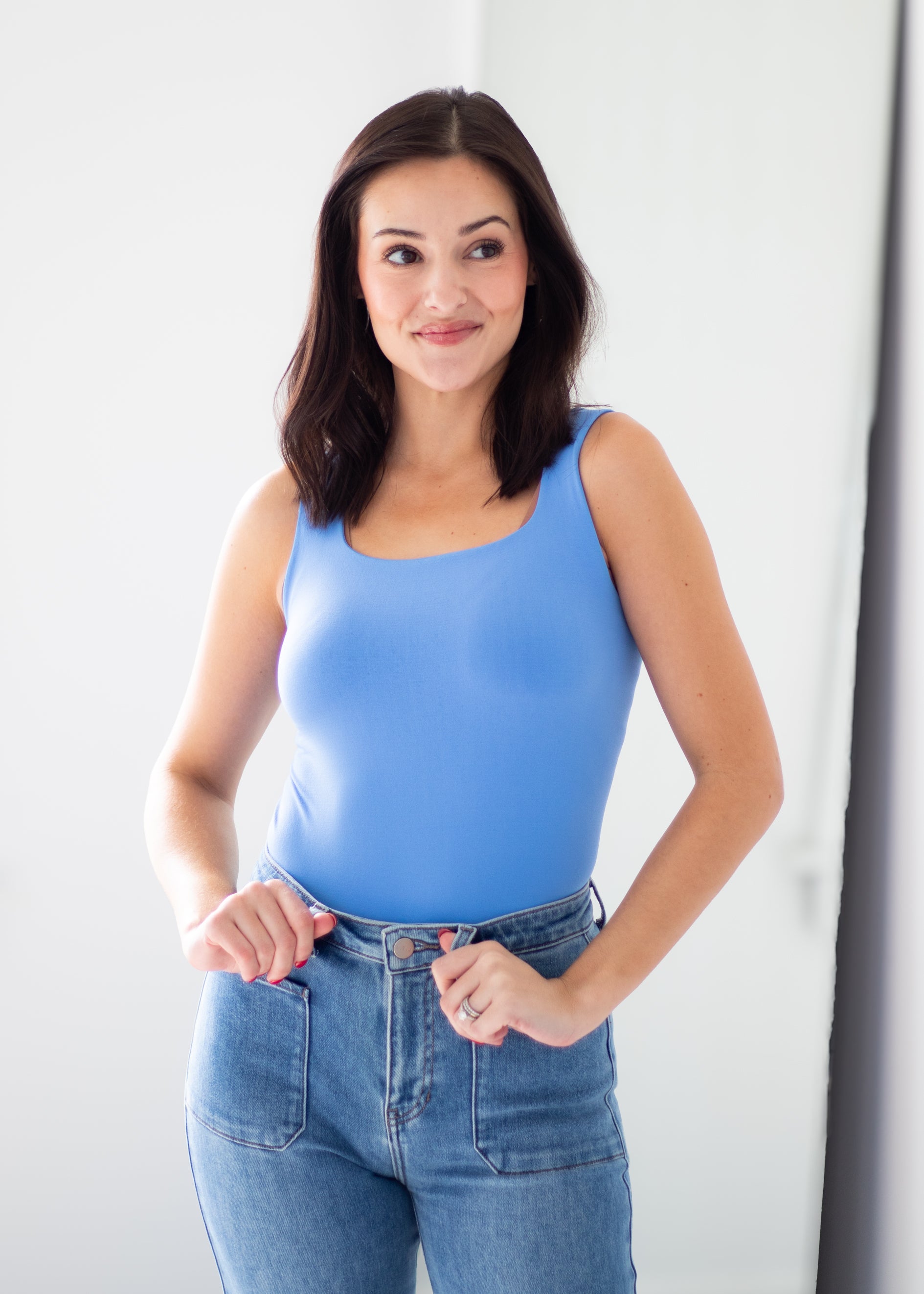 A woman with dark brown hair wearing a bright blue sleeveless scoop-neck tank top and blue jeans, standing against a plain white studio background.