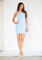 A woman modeling a light blue halter mini dress with white piping and scalloped edges, paired with white heeled sandals, set against a plain white studio background.