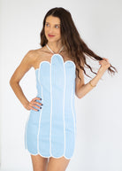 A woman modeling a light blue halter mini dress with vertical white piping and scalloped edges, posed against a plain white studio background.