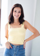 A smiling woman with dark brown hair wearing a pale yellow ribbed tank top and blue jeans, standing in a brightly lit indoor setting.