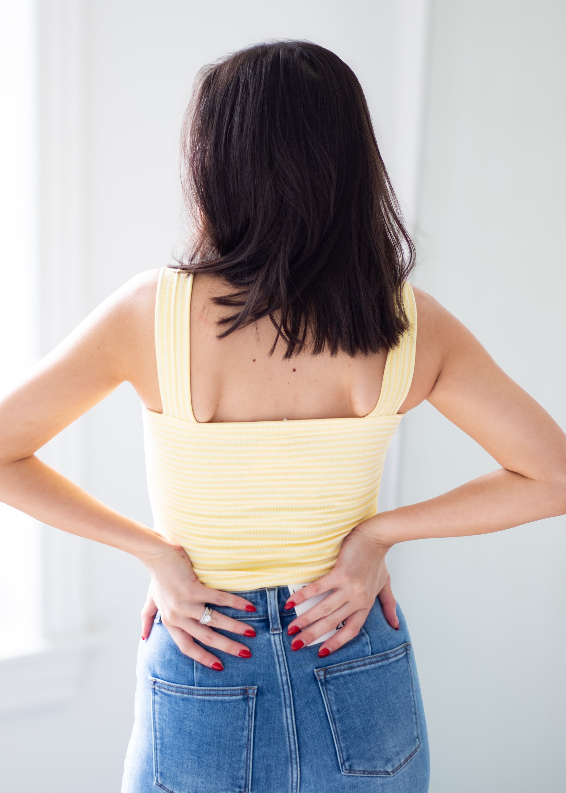 A woman viewed from behind wearing a pale yellow ribbed tank top and blue jeans, standing indoors with her hands on her lower back.