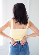 A woman viewed from behind wearing a pale yellow ribbed tank top and blue jeans, standing indoors with her hands on her lower back.