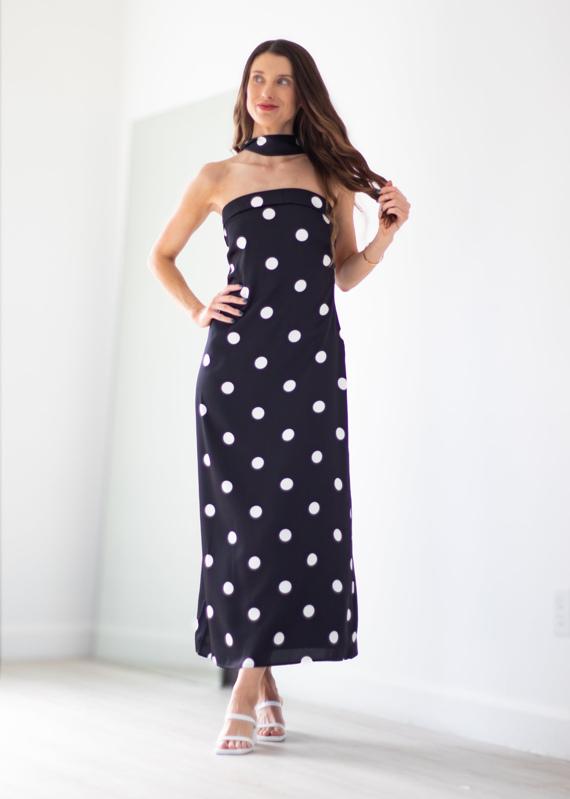 A woman modeling a strapless black halter maxi dress with large white polka dots, standing in a bright white room.