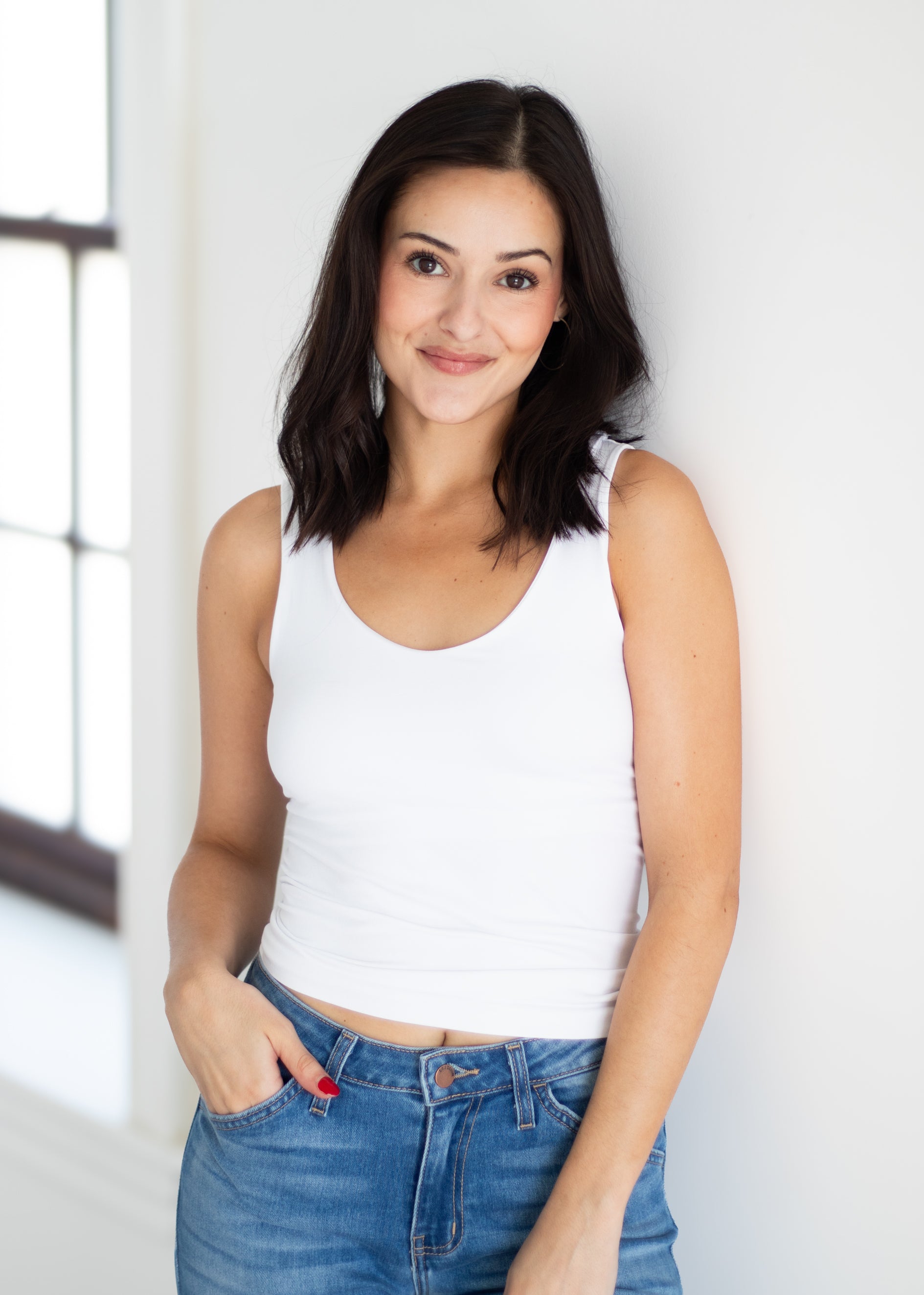 A woman with dark shoulder-length hair wearing a white ribbed tank top and blue jeans, standing against a white wall near a window.