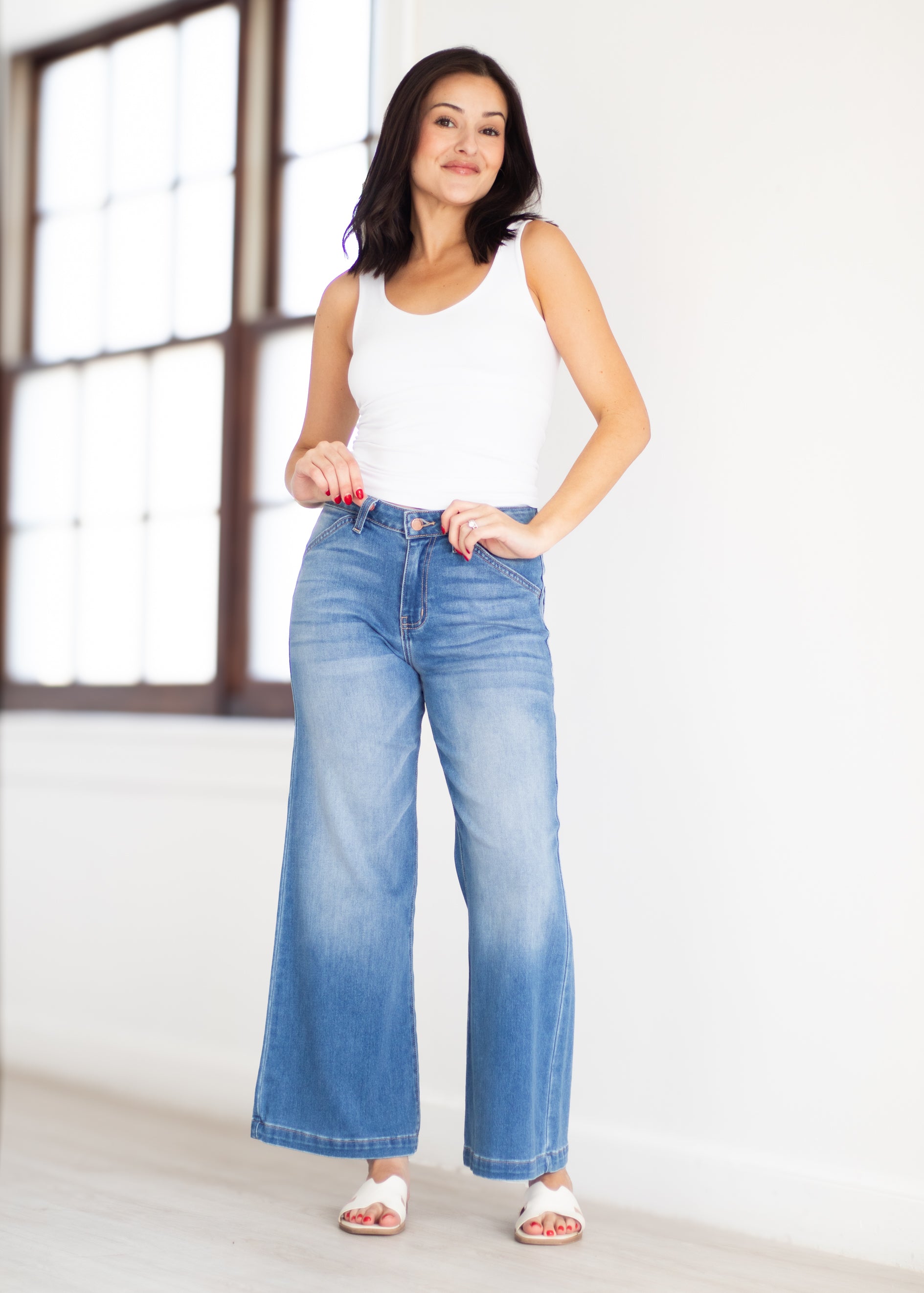 A woman modeling a pair of wide-leg light blue denim jeans with a faded wash, paired with a white tank top and white slide sandals, standing in a bright room with a window.
