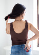 A woman shown from behind wearing a brown ribbed tank top with lace trim on the straps and blue jeans, standing in a bright indoor setting.