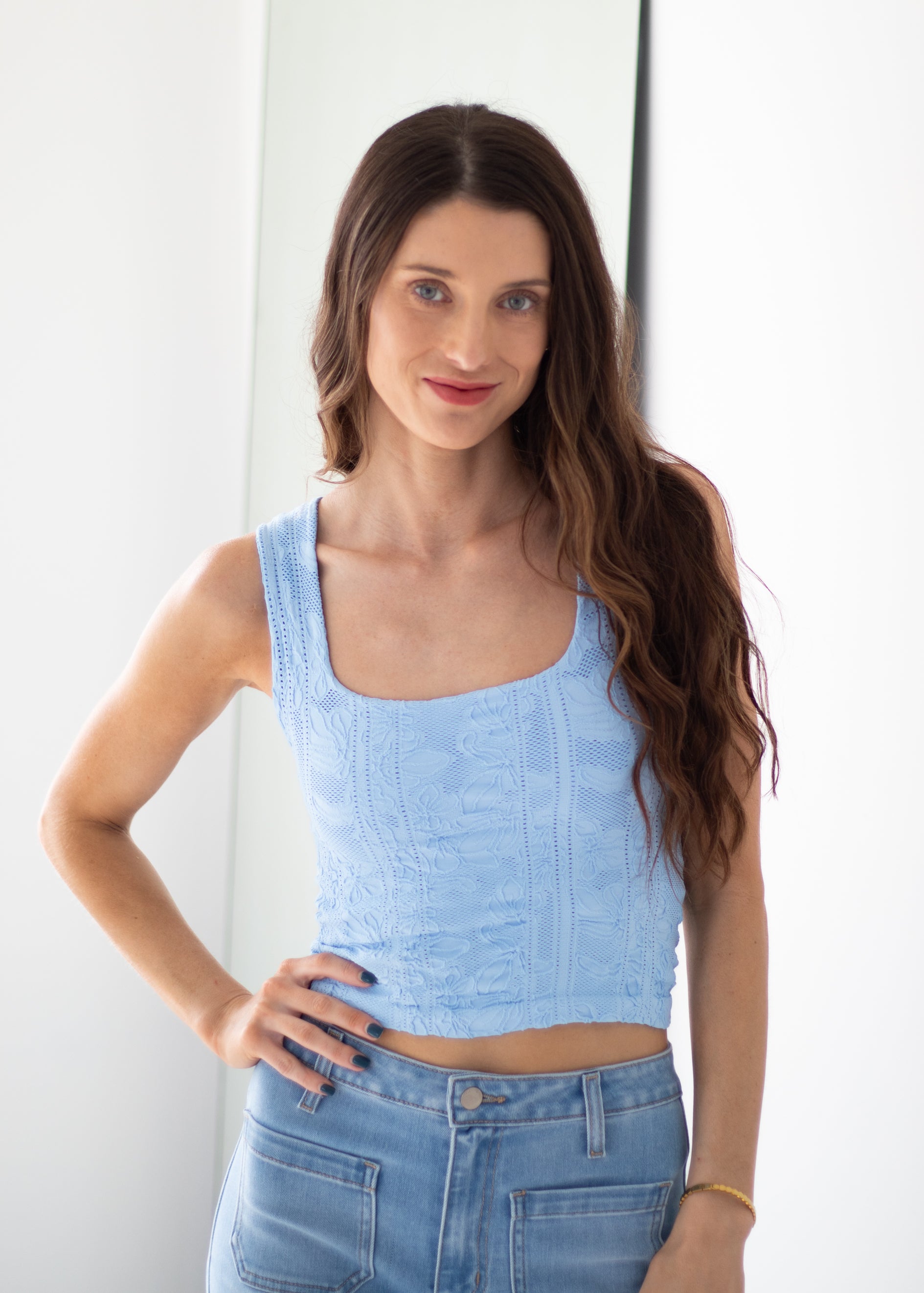 A woman with long brown hair wearing a light blue textured sleeveless crop top and light blue jeans, standing against a white wall.