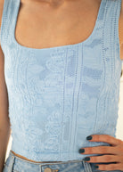 A close-up of a person wearing a light blue textured knit tank top with floral lace-like patterns, paired with blue jeans.