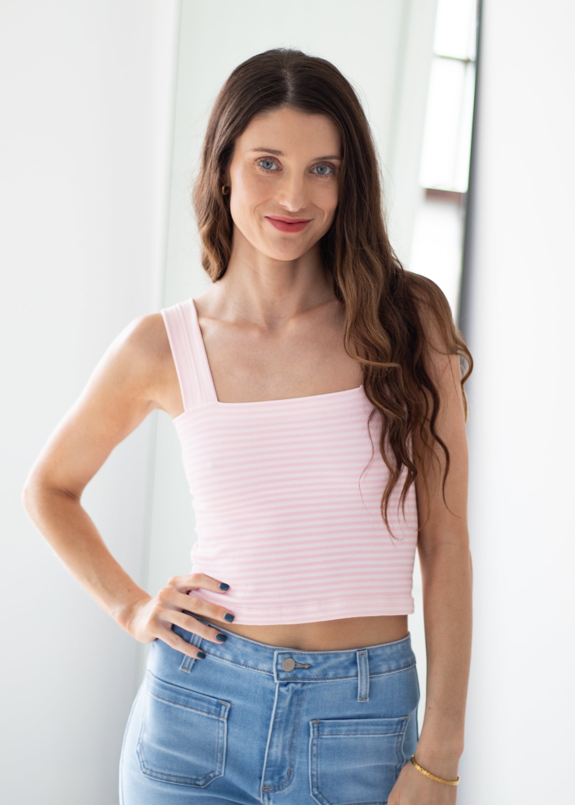A woman with long brown hair wearing a light pink and white horizontally striped ribbed tank top and light blue jeans, posing in a brightly lit indoor setting.